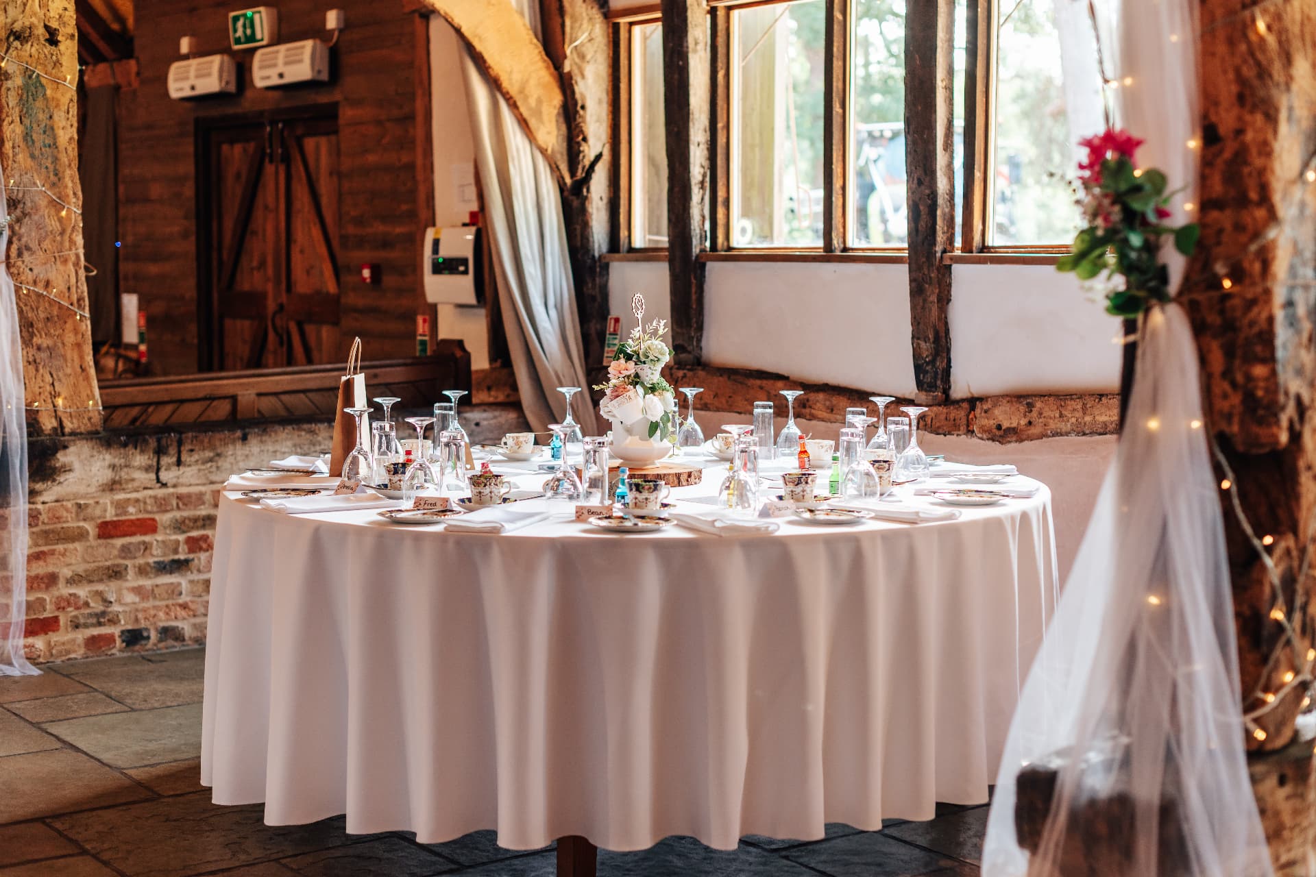 Round barn wedding table laid with white linen, vintage china teacups and glassware beneath fairy lights