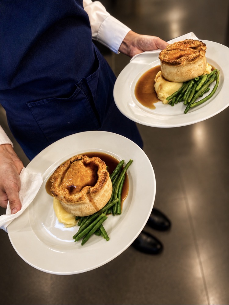 Server carrying two golden pastry pies with mash, green beans and rich gravy