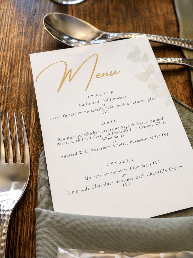 Gold-script wedding menu card on a sage napkin with hammered cutlery on a wooden table