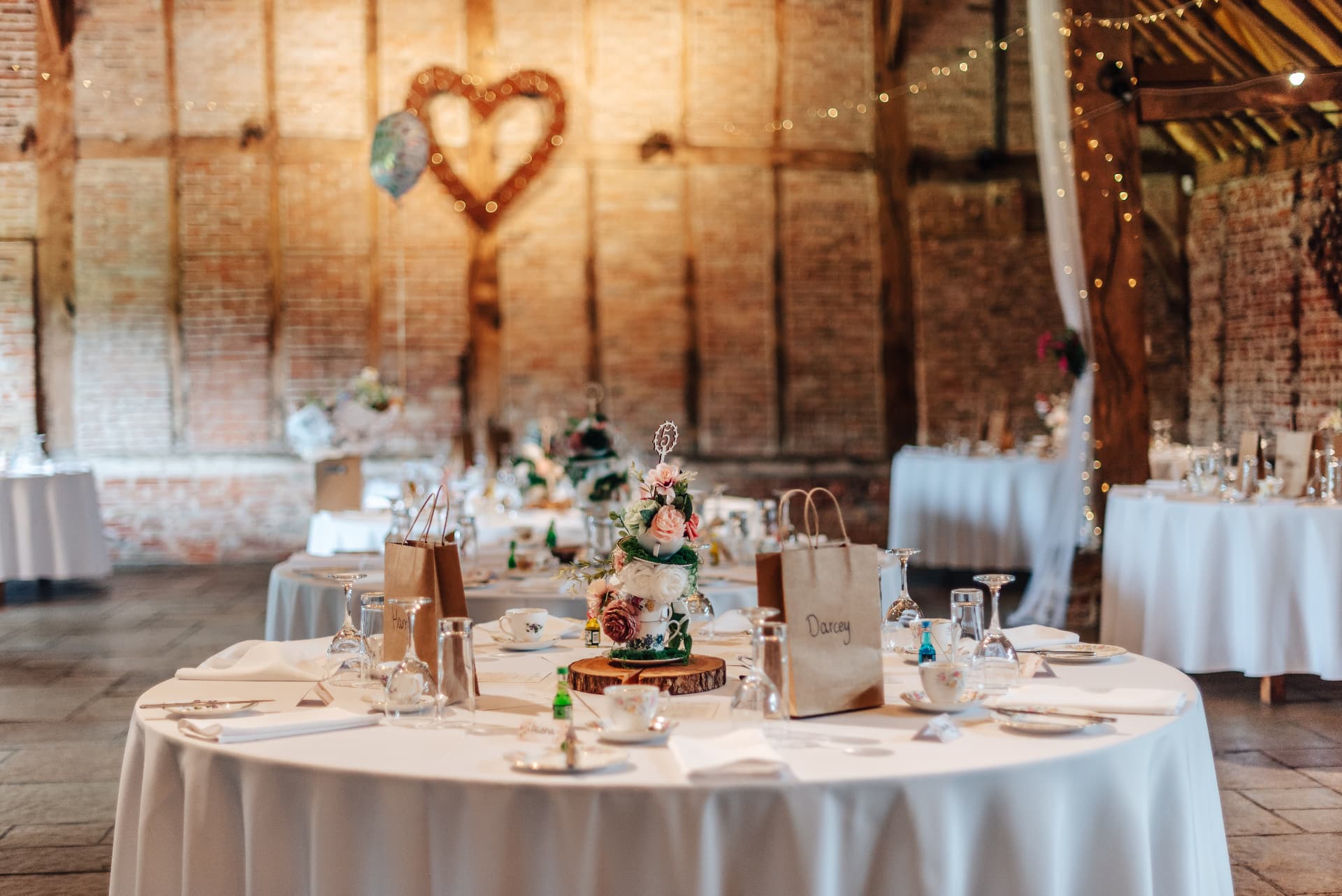 Barn wedding breakfast tables dressed in white linen with vintage teacups and a wicker heart on the brick wall