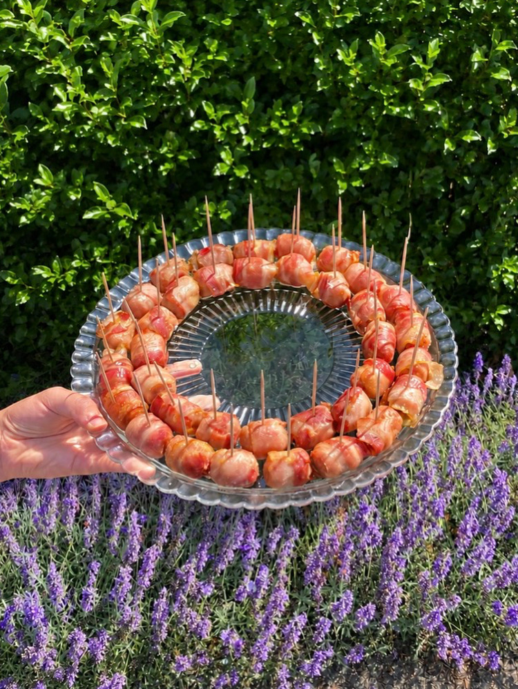 Bacon-wrapped chipolatas on a glass platter held in front of a lavender border